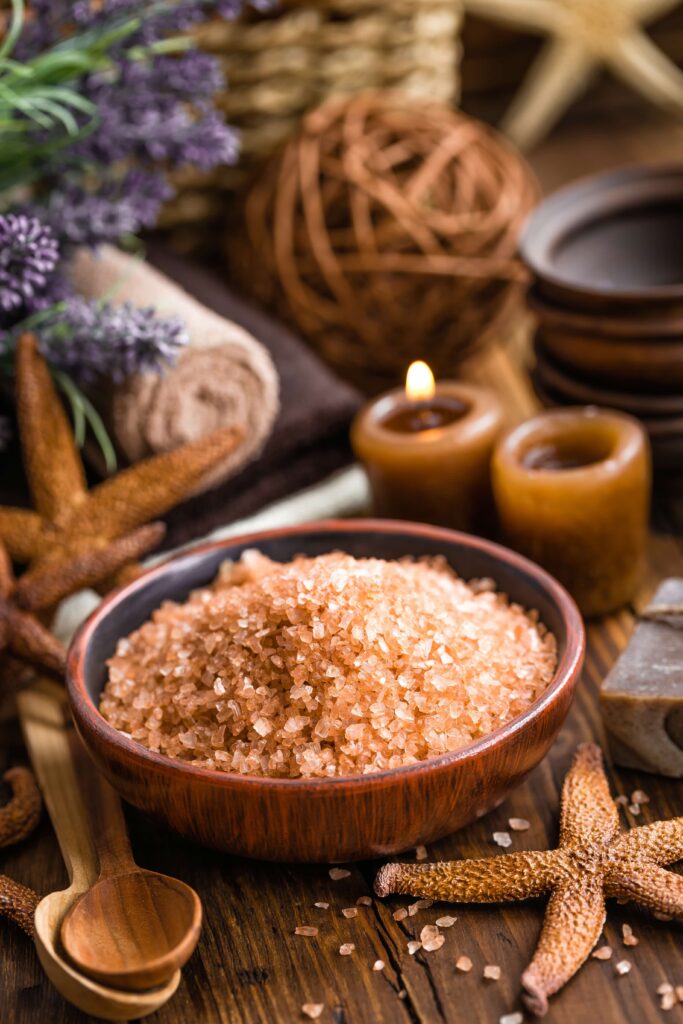 A wooden bowl filled with pink salts sits on a rustic table, surrounded by starfish, candles, a wooden spoon, towels, and decorative spa items.